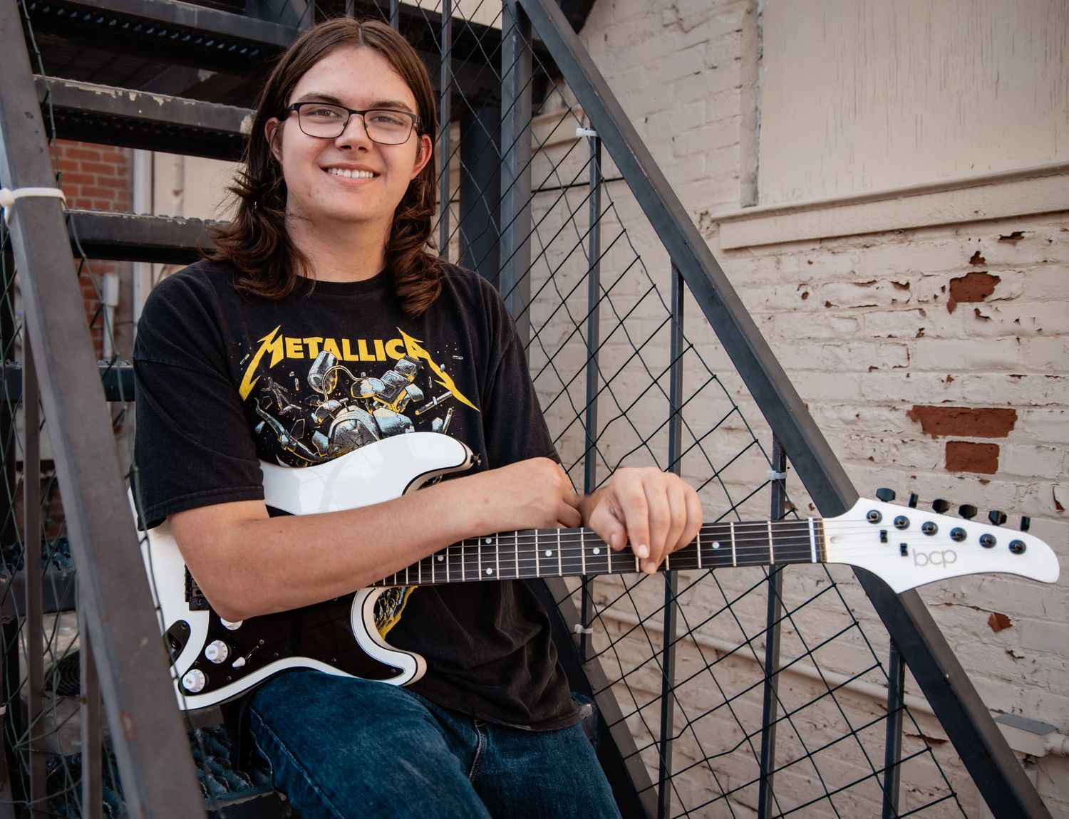 Young musician in Metallica shirt smiling while holding electric guitar on metal staircase outdoors.
