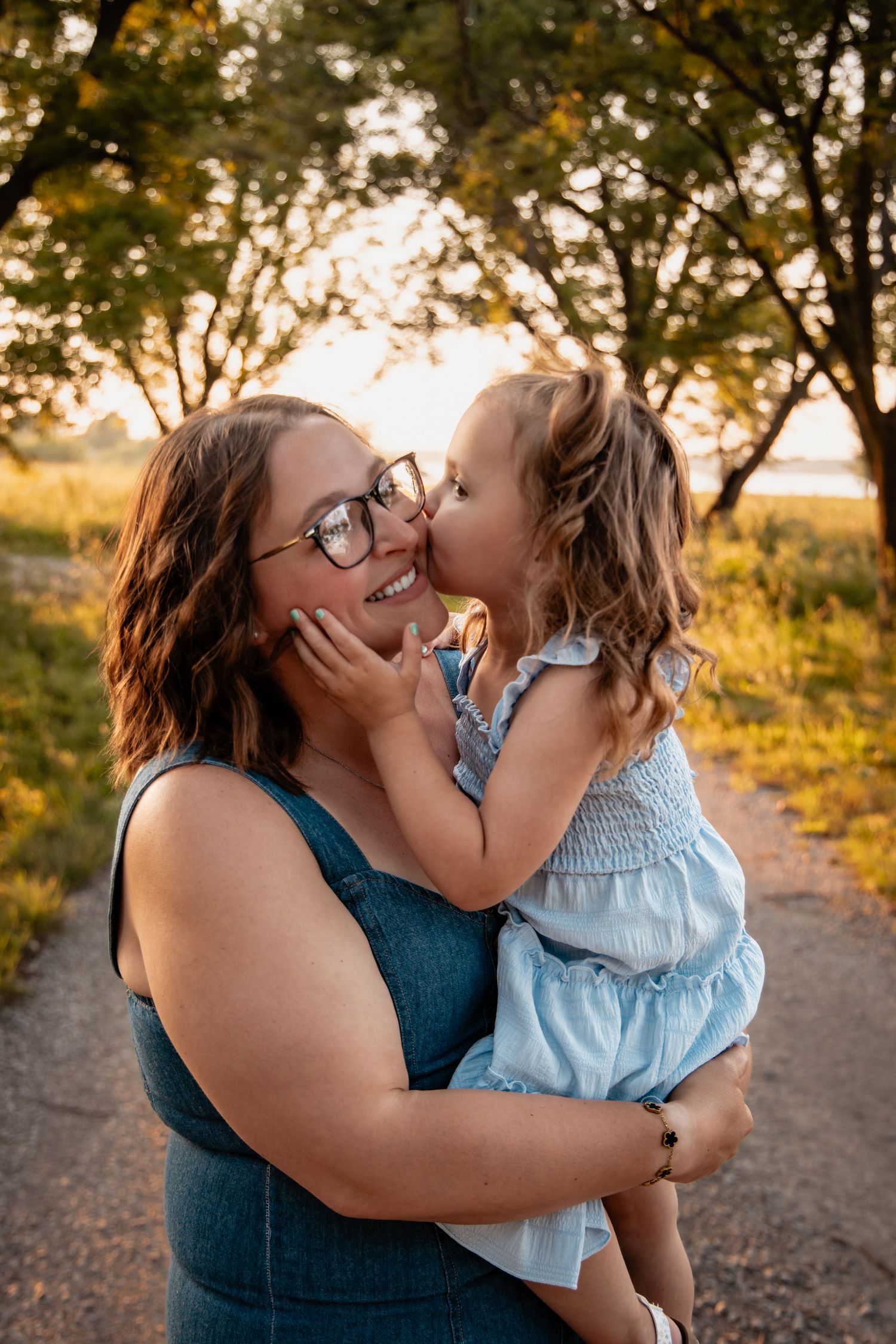 A woman in glasses holds a young girl who kisses her cheek outdoors at golden hour.