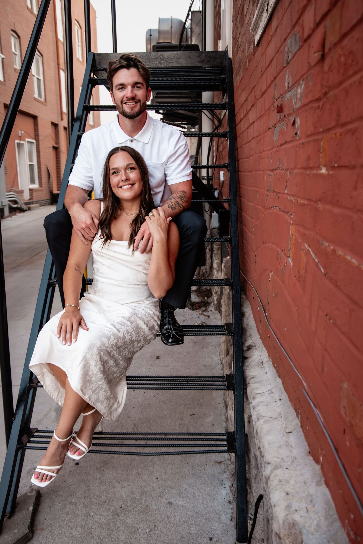 Smiling couple in wedding attire posing on urban fire escape stairs beside red brick wall.