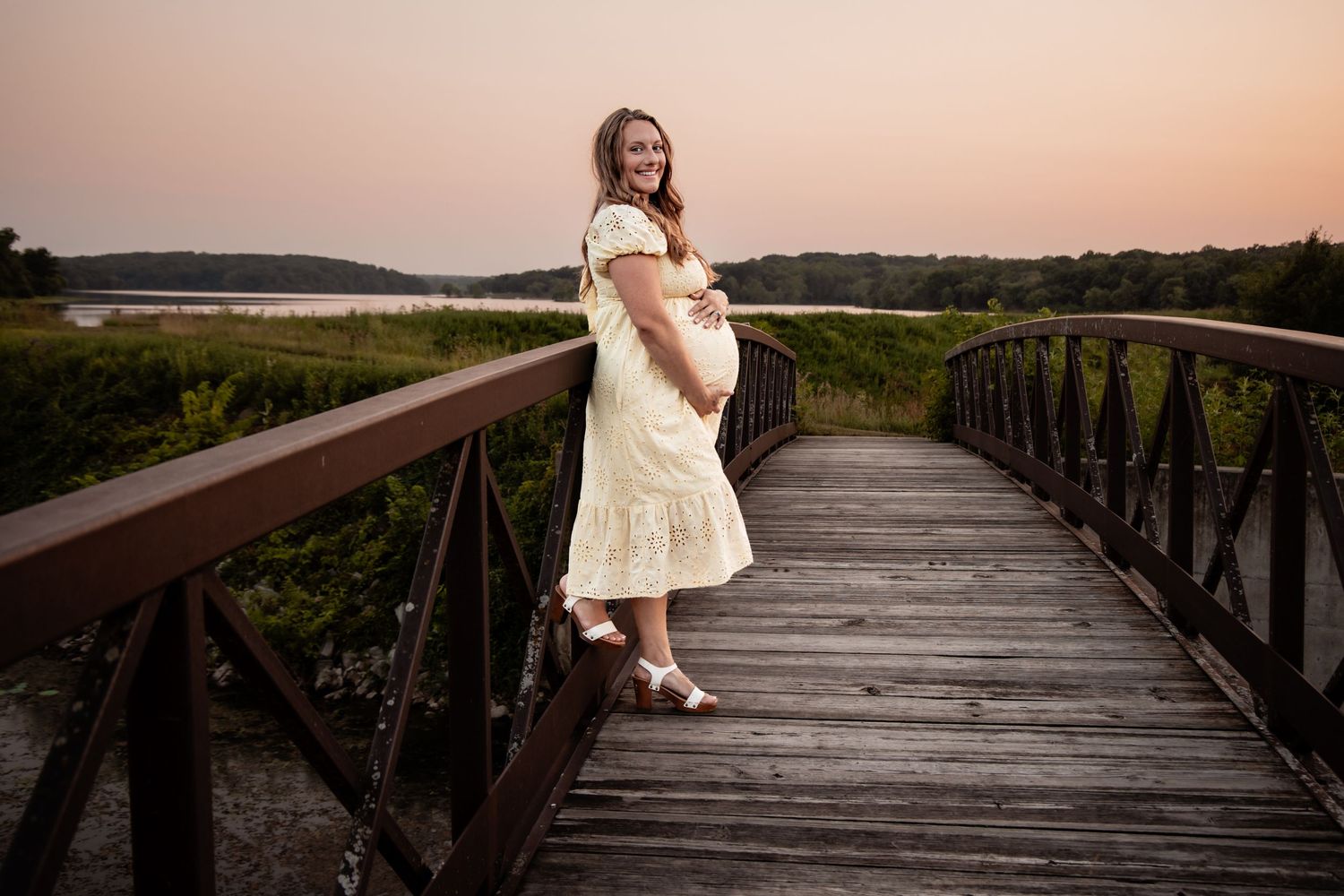 Pregnant woman in white dress on wooden bridge at sunset with scenic water and greenery backdrop.