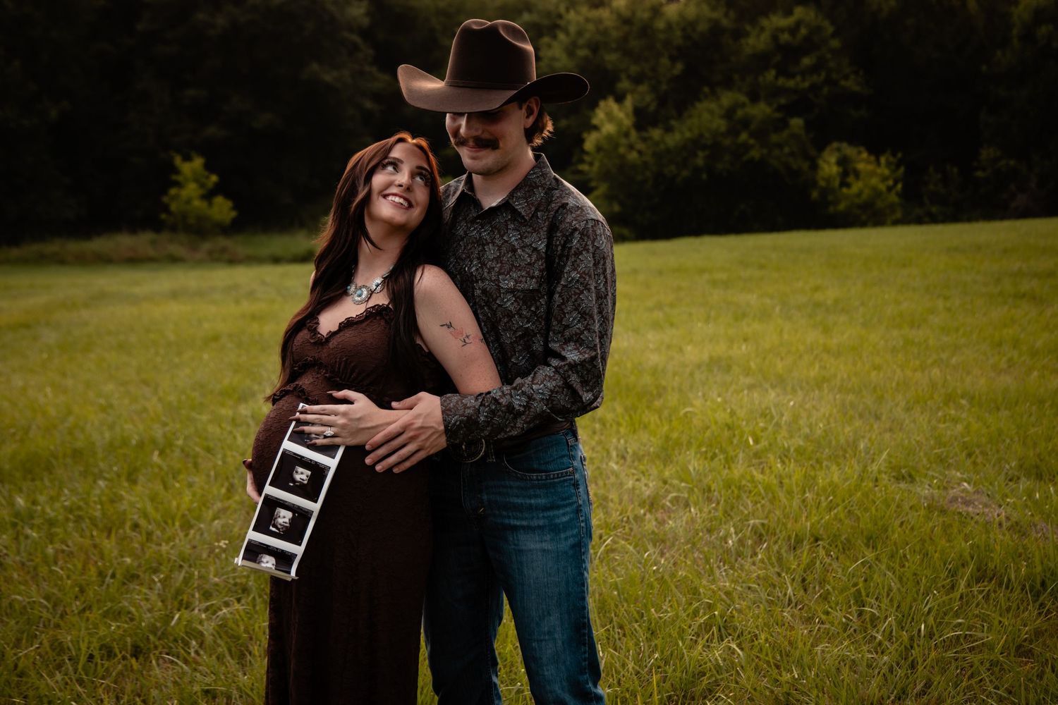 Pregnant couple in a field holding ultrasound photos, woman in burgundy dress, man in cowboy hat and floral shirt.