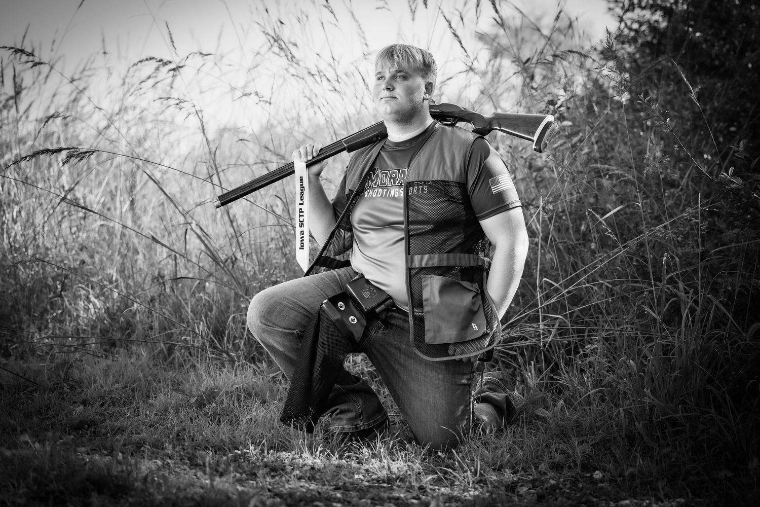 Black and white photo of a hunter kneeling outdoors with a shotgun over his shoulder in a field.