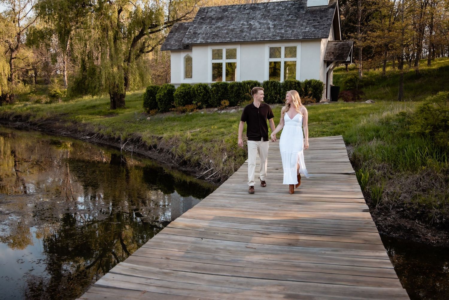 An engaged couple walks hand in hand along a wooden boardwalk beside a pond near a white cottage.