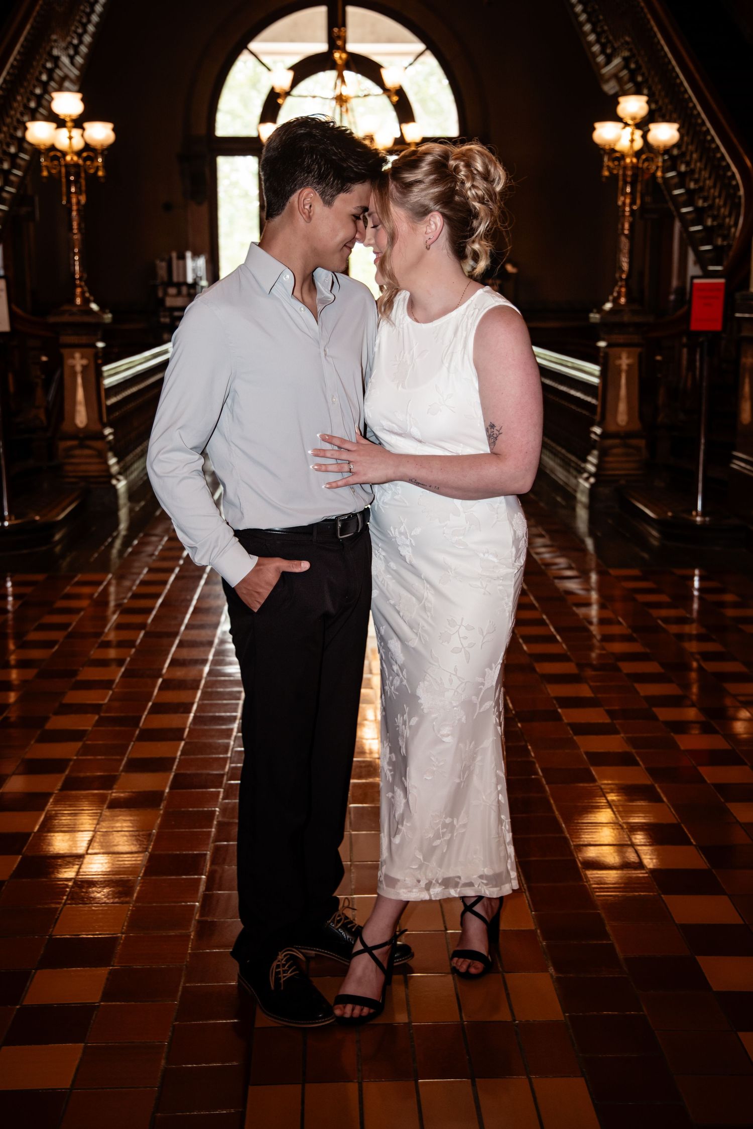 Couple in wedding attire sharing a romantic moment in an elegant hall with ornate chandeliers and checkered tile floors.