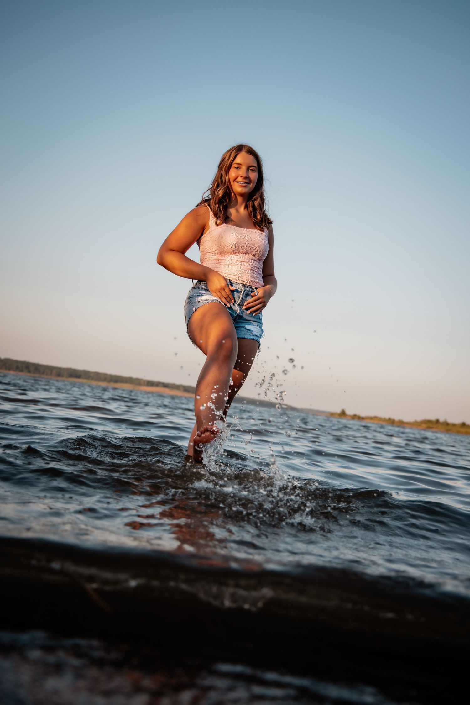 Young woman in white top and denim shorts splashing water at the beach during golden hour sunset.