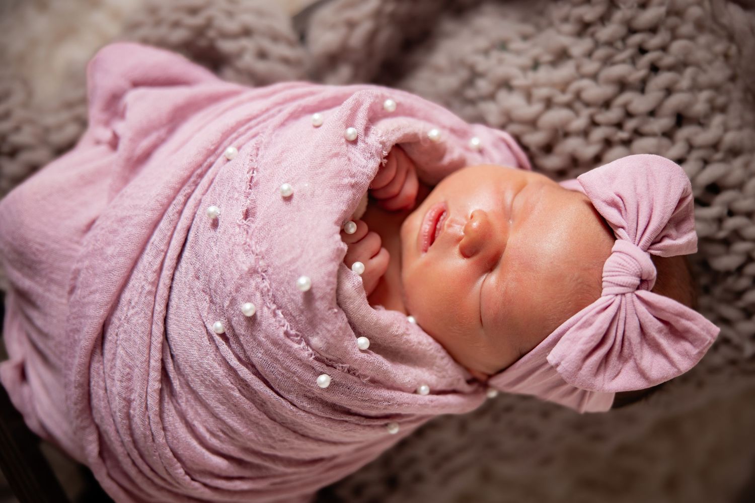 Newborn baby girl sleeping peacefully wrapped in a pink swaddle blanket with pearl accents and matching bow headband.