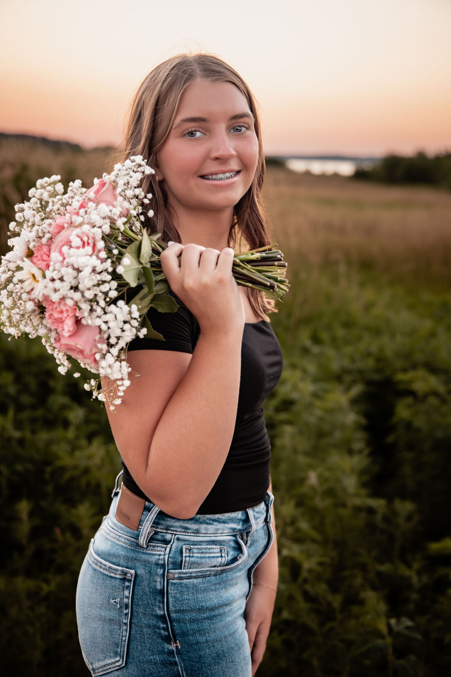 Smiling teenage girl holding pink and white flower bouquet in a field at sunset wearing black top and jeans.