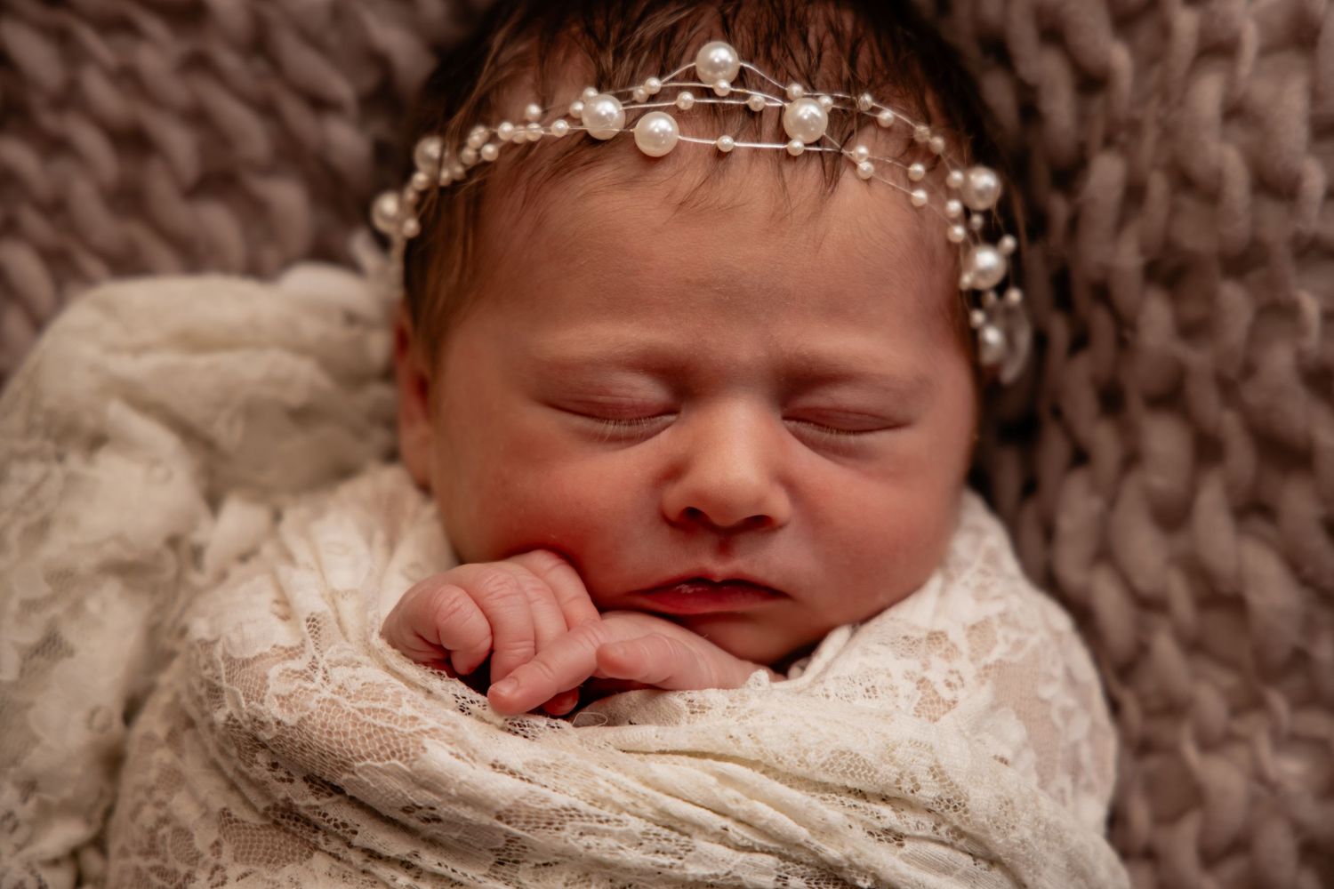 Peaceful newborn baby girl sleeping wrapped in white lace swaddle wearing delicate pearl and crystal headband.