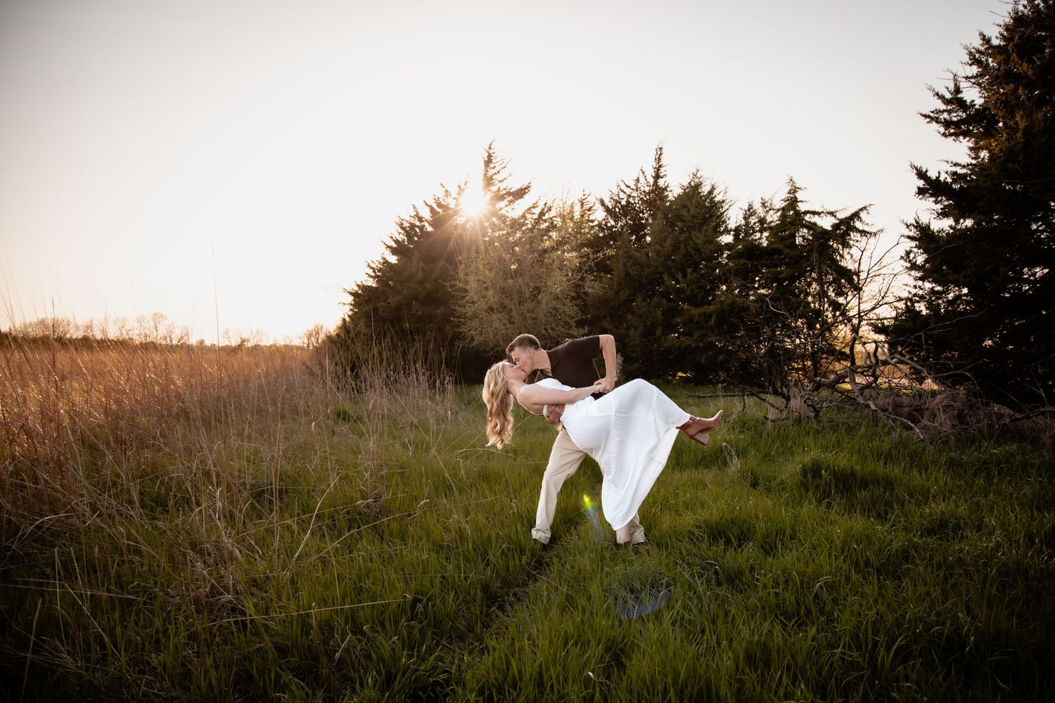 A couple shares a romantic dip kiss in a sunlit meadow surrounded by tall golden grass and evergreen trees.