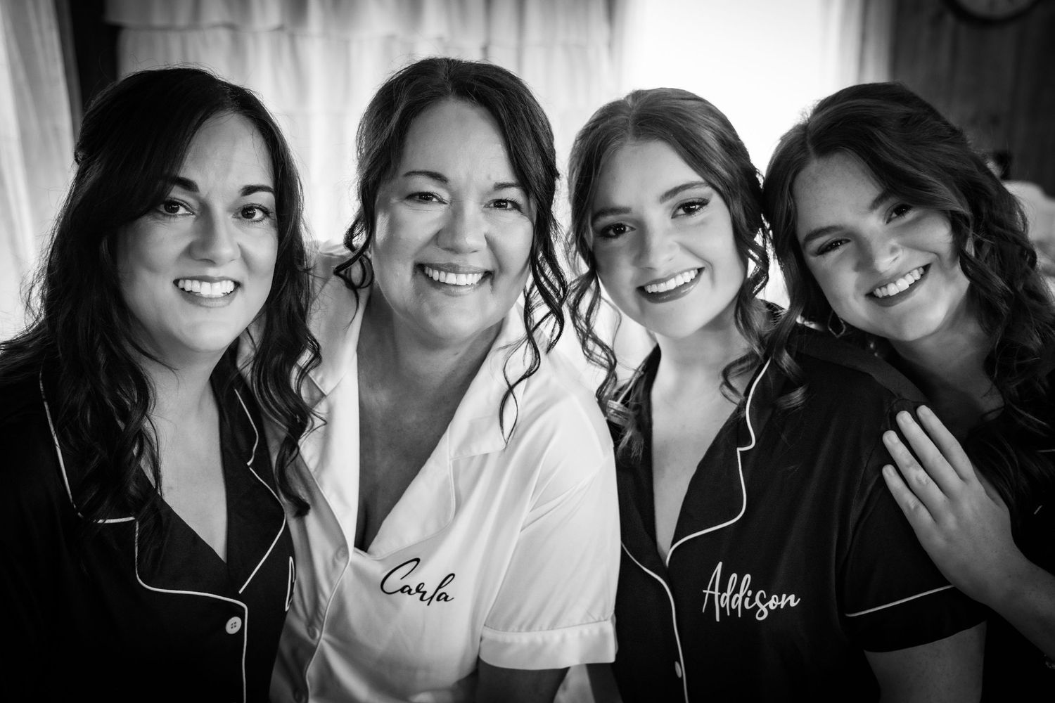 Black and white photo of smiling bride in white robe with three bridesmaids in black robes getting ready together.