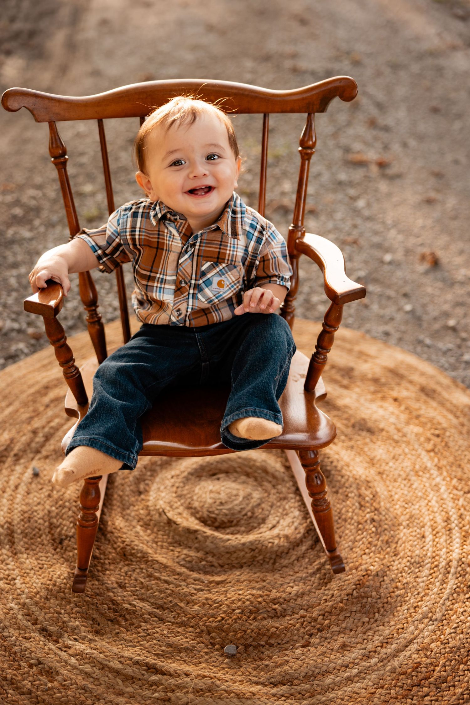 Smiling baby boy in plaid shirt and jeans sitting in a wooden rocking chair on a round braided rug outdoors.