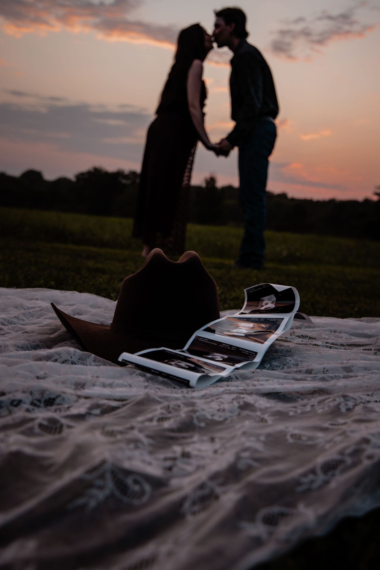 Couple kissing at sunset silhouette with cowboy hat and photo strip on lace blanket in foreground.