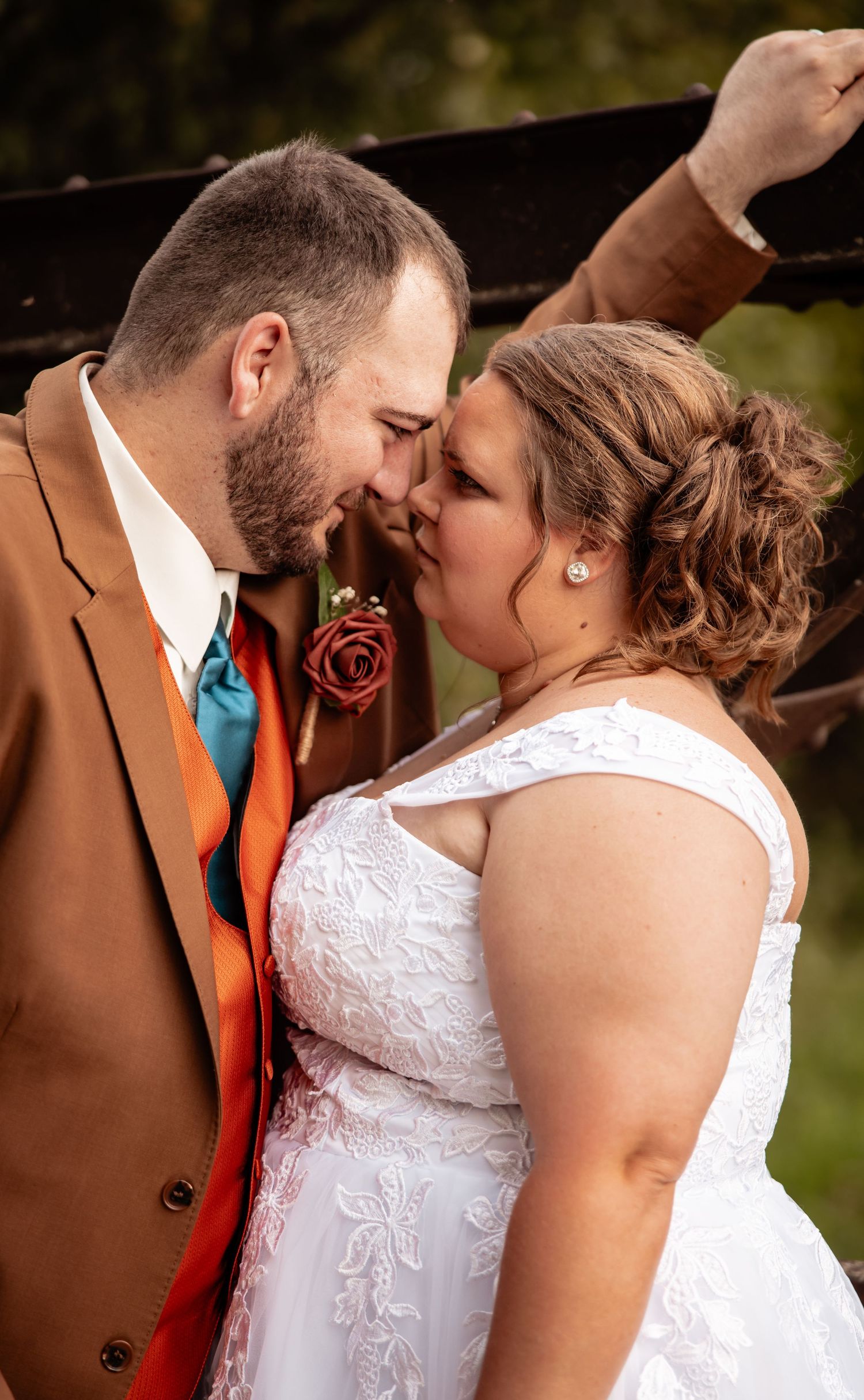 Bride and groom share a romantic nose-to-nose moment, groom in brown suit with teal and orange tie, bride in white lace gown.