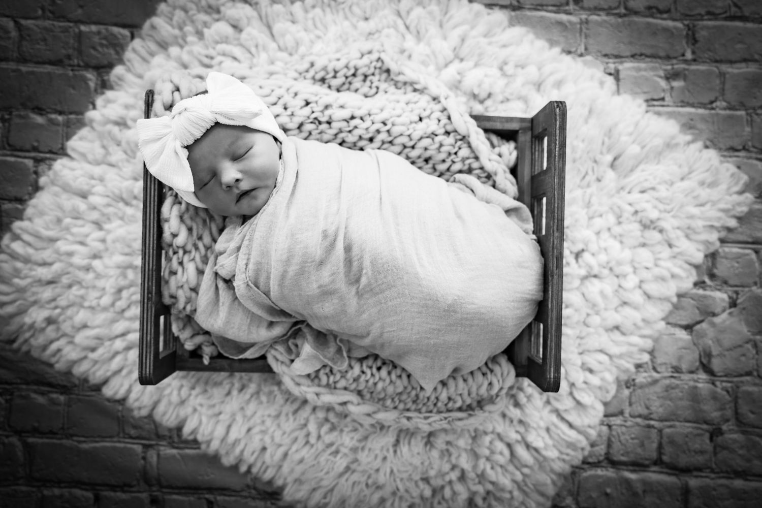 Black and white photo of swaddled newborn with bow headband sleeping in wooden mini crib on fluffy rug.