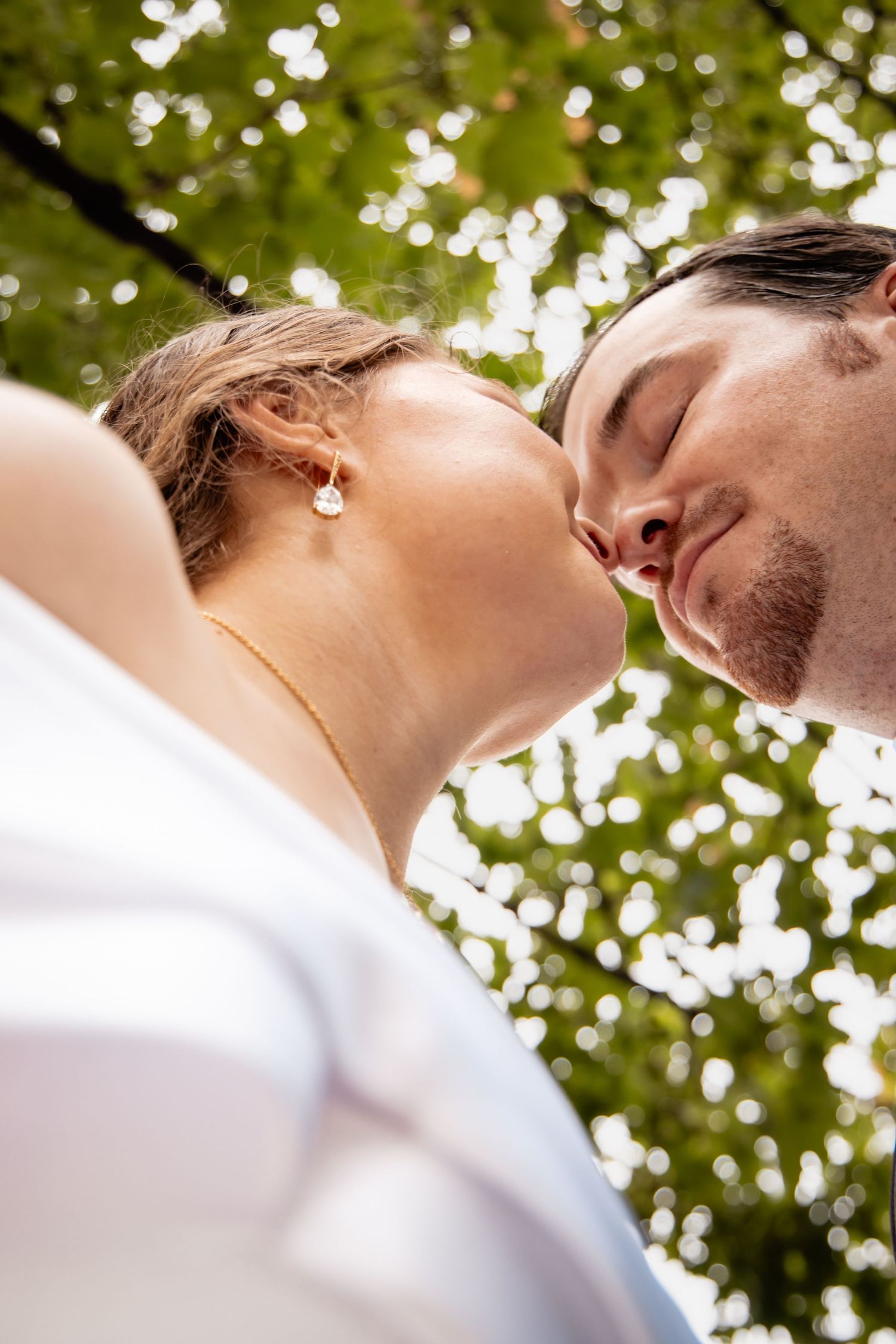 Low-angle view of a romantic couple about to kiss outdoors under green tree canopy on a sunny day.
