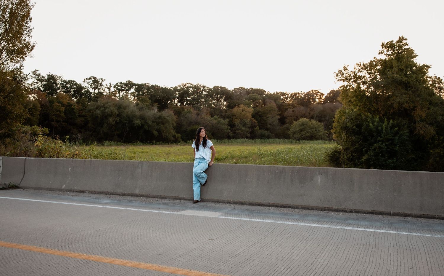 A woman in light blue scrubs leans against a highway barrier with green trees and fields in the background.