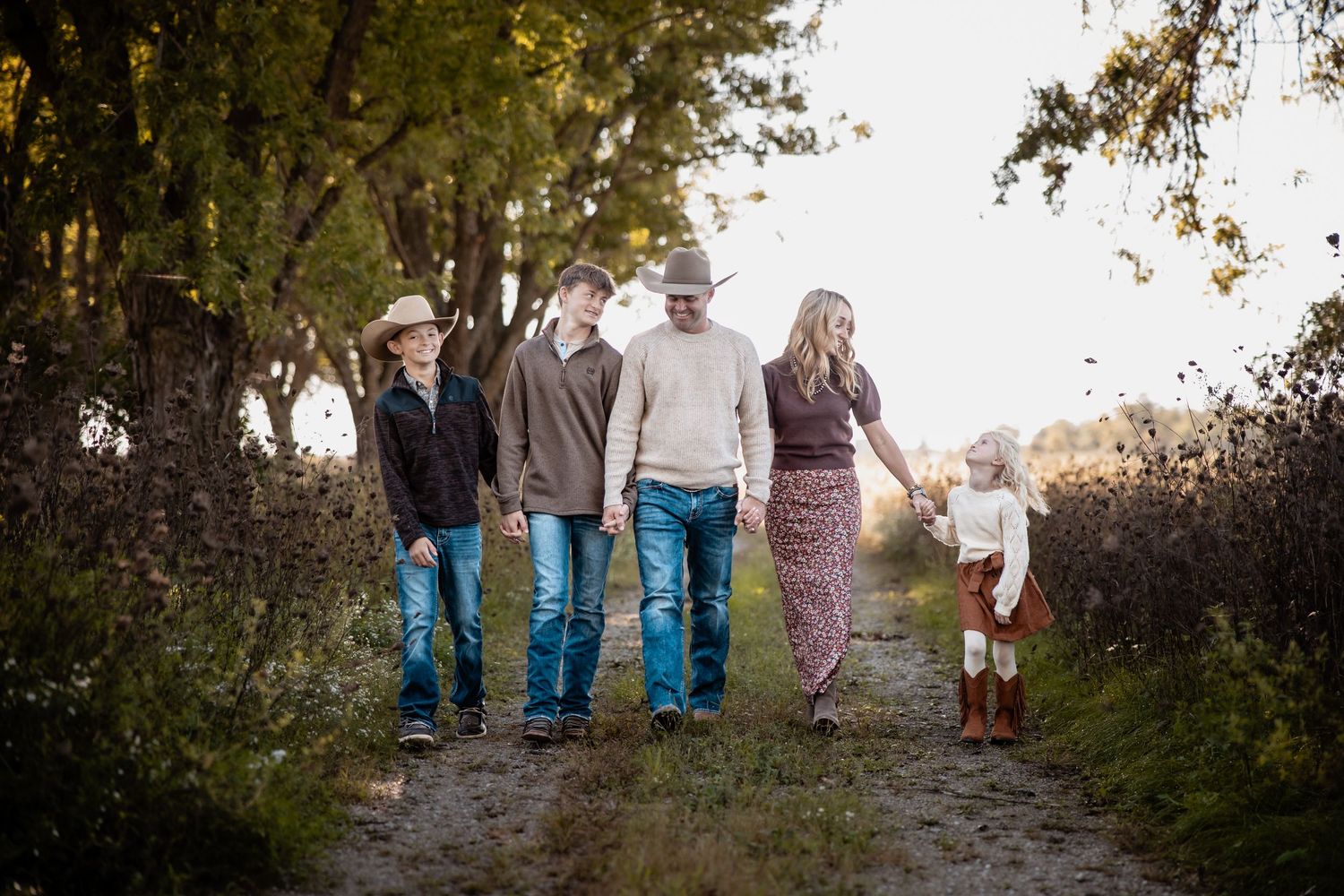 Family of five walking down a rustic dirt path surrounded by autumn trees in a warm outdoor photo session.