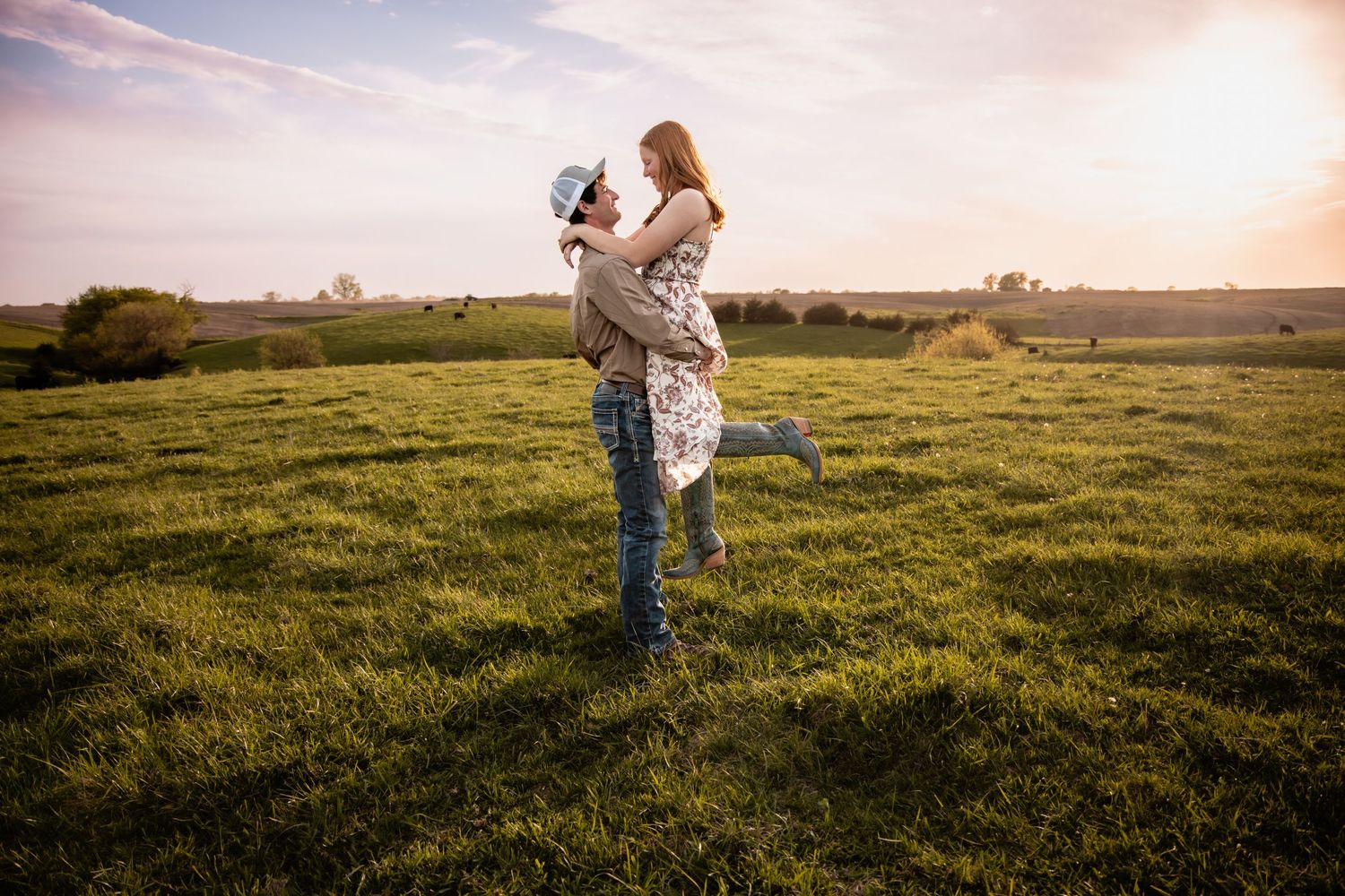 A man lifts a laughing woman in a floral dress in a lush green field during a glowing sunset.