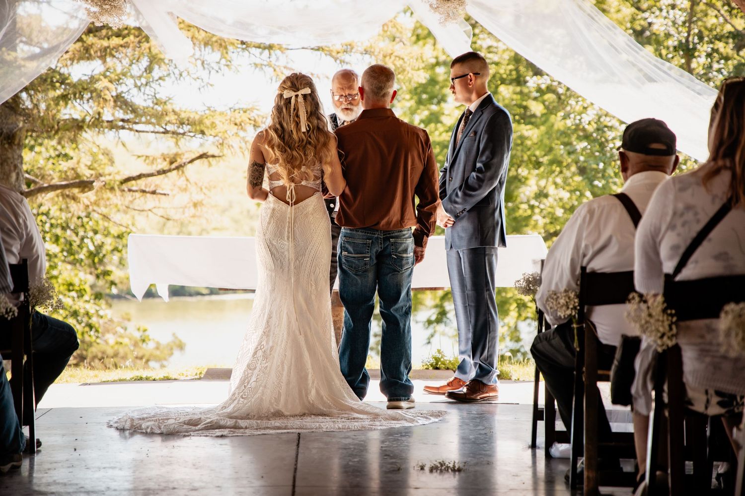 Outdoor wedding ceremony with bride in lace gown, groom in suit, and officiant under draped canopy near water.