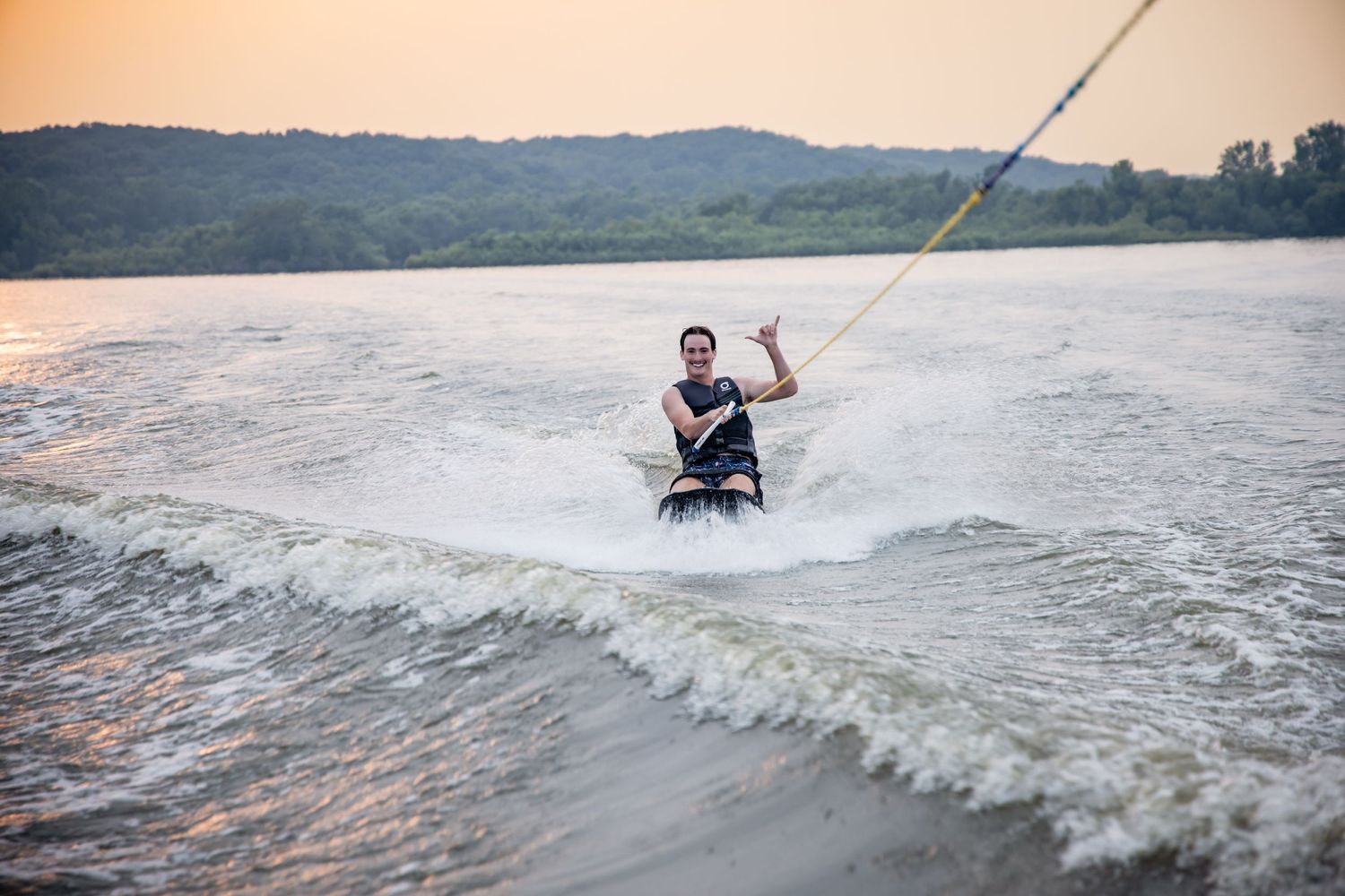 Person wakeboarding on a lake at sunset, waving at the camera while being towed by a boat.