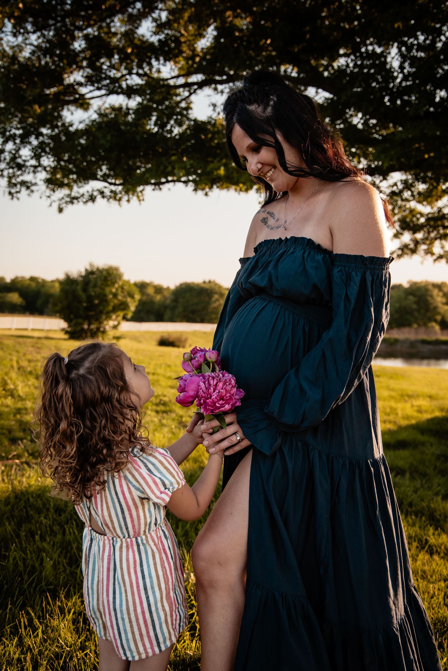 A young girl offers pink flowers to her pregnant mother in a teal gown at golden hour.