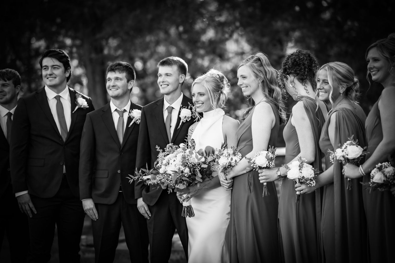 Black and white wedding party photo with bride, groom, groomsmen, and bridesmaids holding bouquets outdoors.