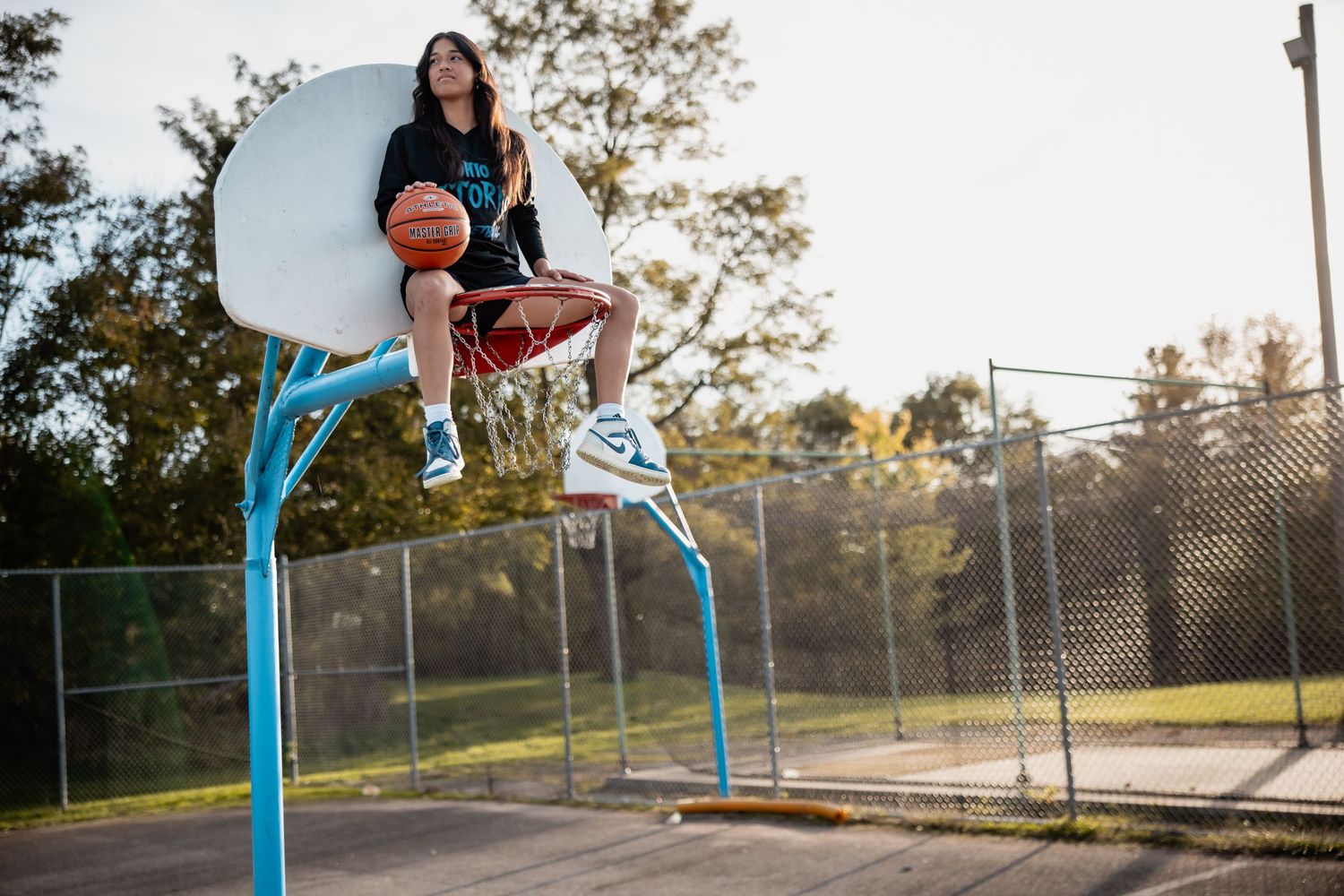 A young woman sits on a basketball hoop backboard at an outdoor court, holding a ball, wearing sneakers and casual attire.