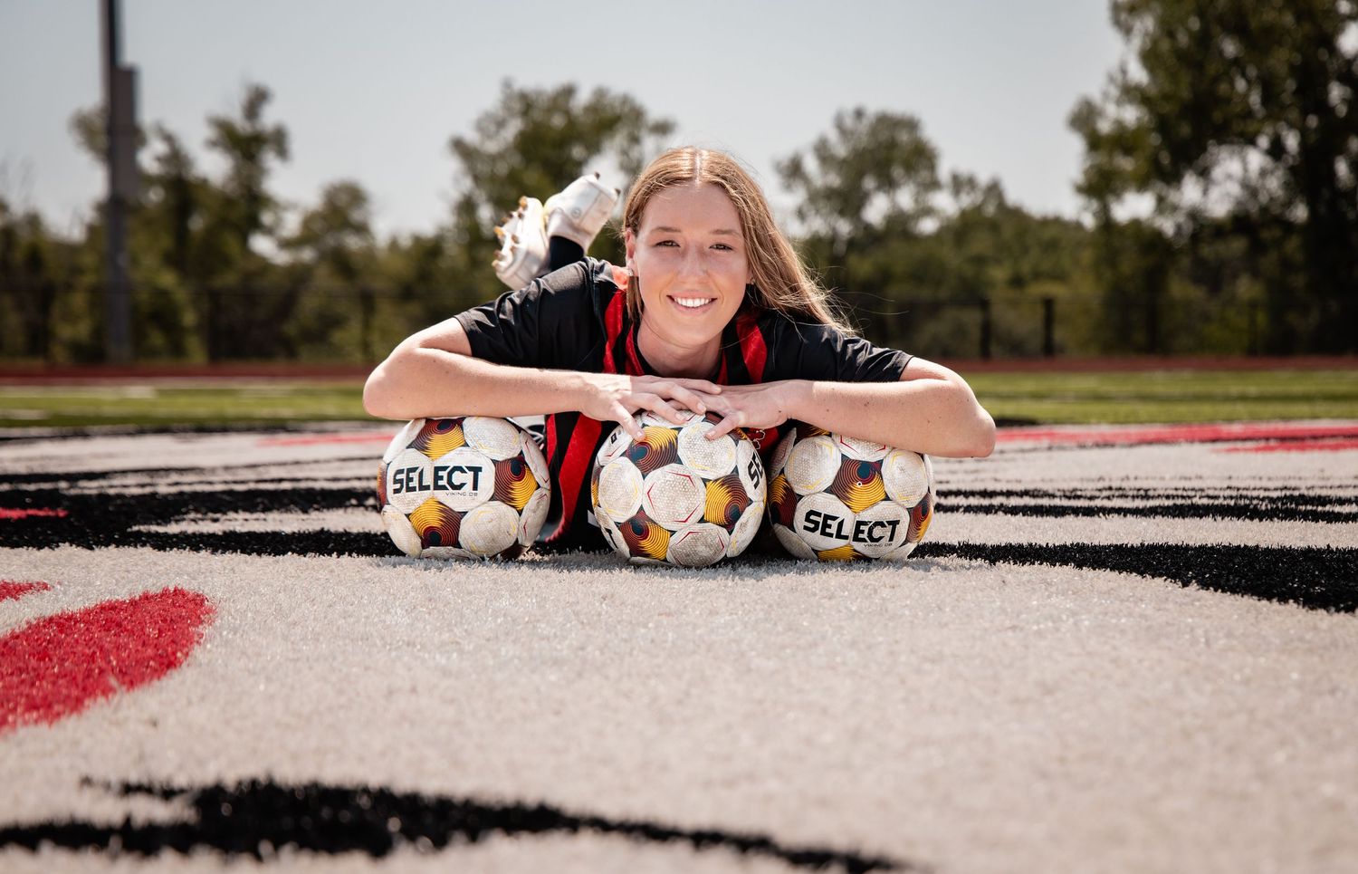Female soccer player posing on field with three Select soccer balls, smiling at camera in black athletic wear.