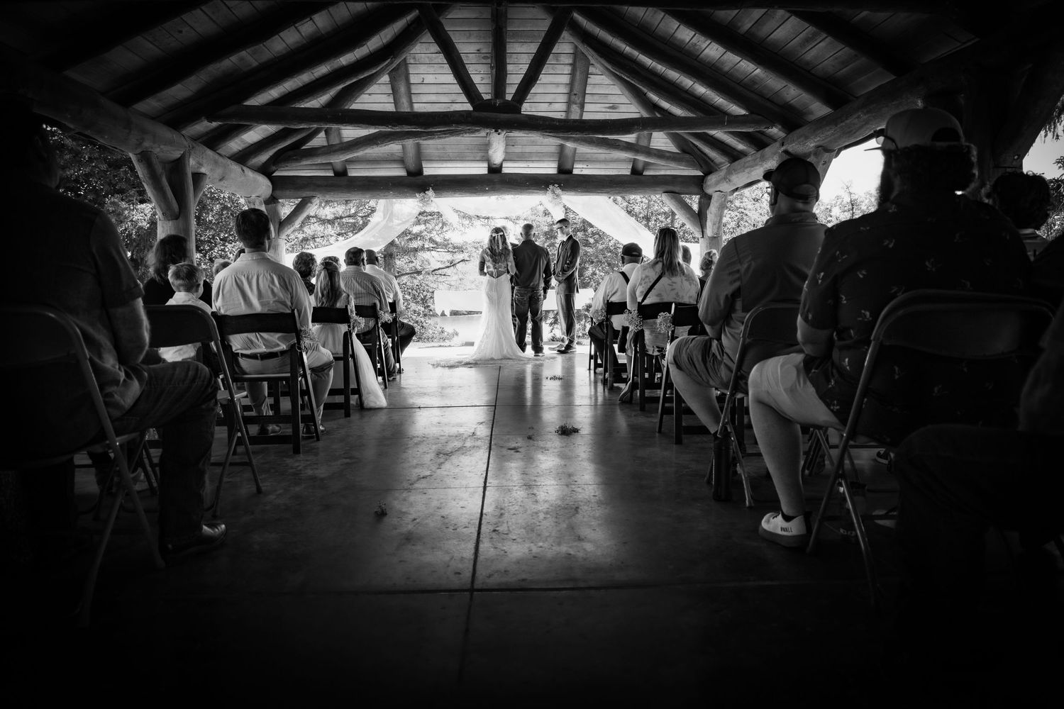 Black and white photo of a wedding ceremony under a rustic pavilion with guests seated in rows facing the couple.