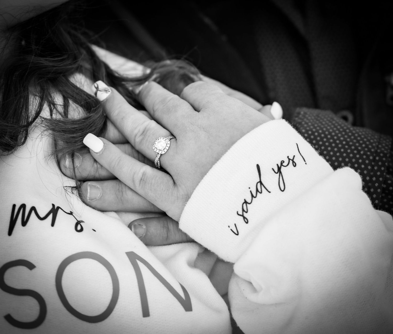 Black and white close-up of engagement ring on hand with Mrs. and I said yes text visible.