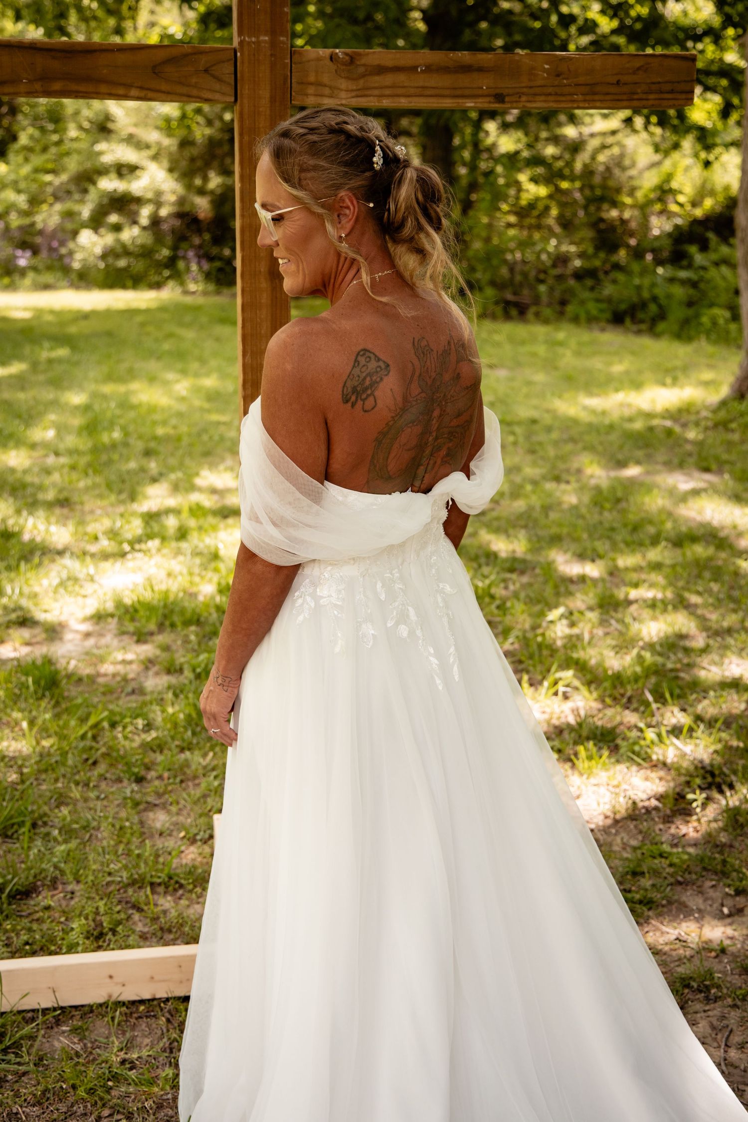 Bride in white off-shoulder gown shows back tattoo while standing near a wooden cross outdoors.