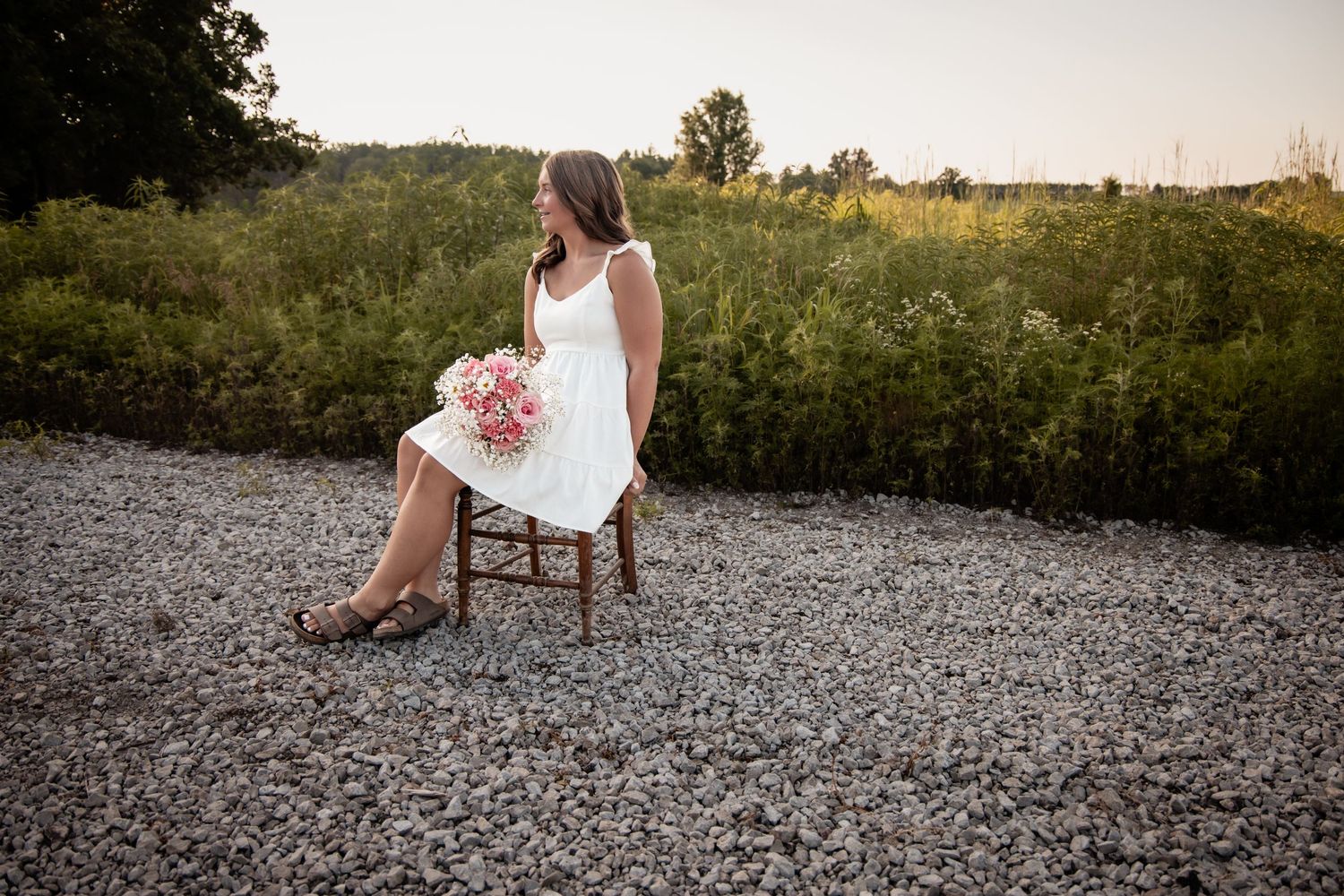 Bride in white dress holding pink bouquet sitting on wooden chair on gravel path surrounded by lush greenery.