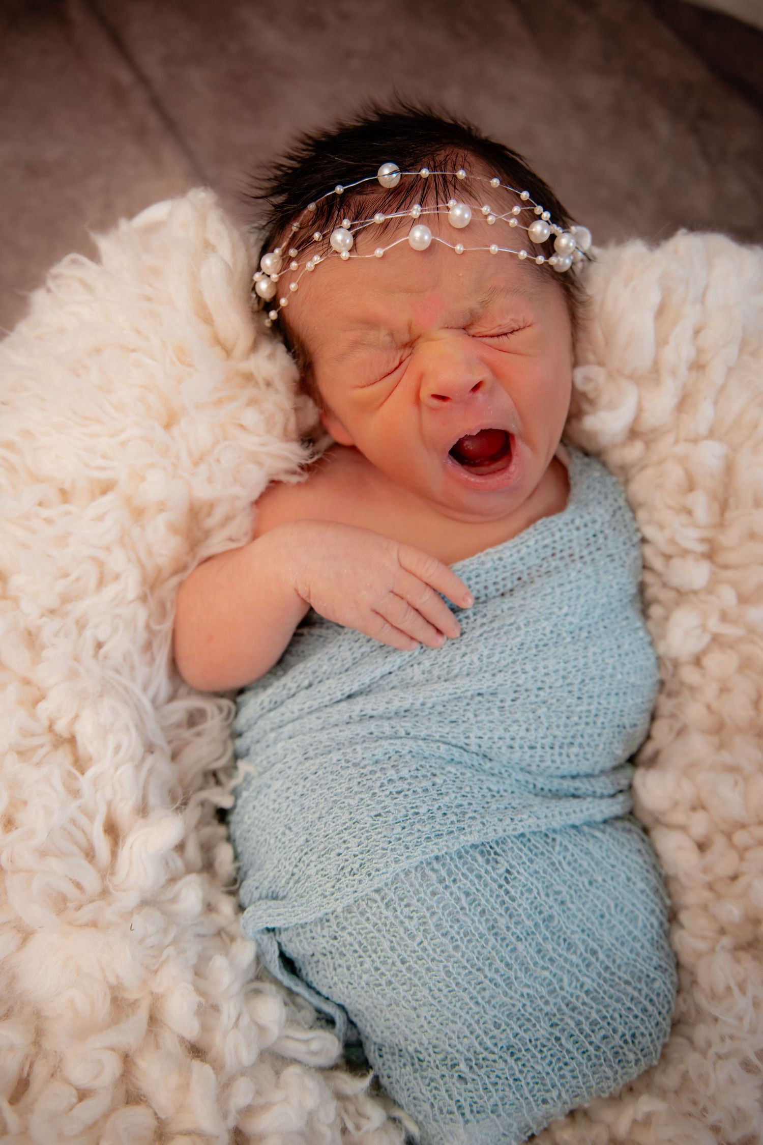 Newborn baby swaddled in blue wrap with floral headband, yawning on fluffy white blanket.
