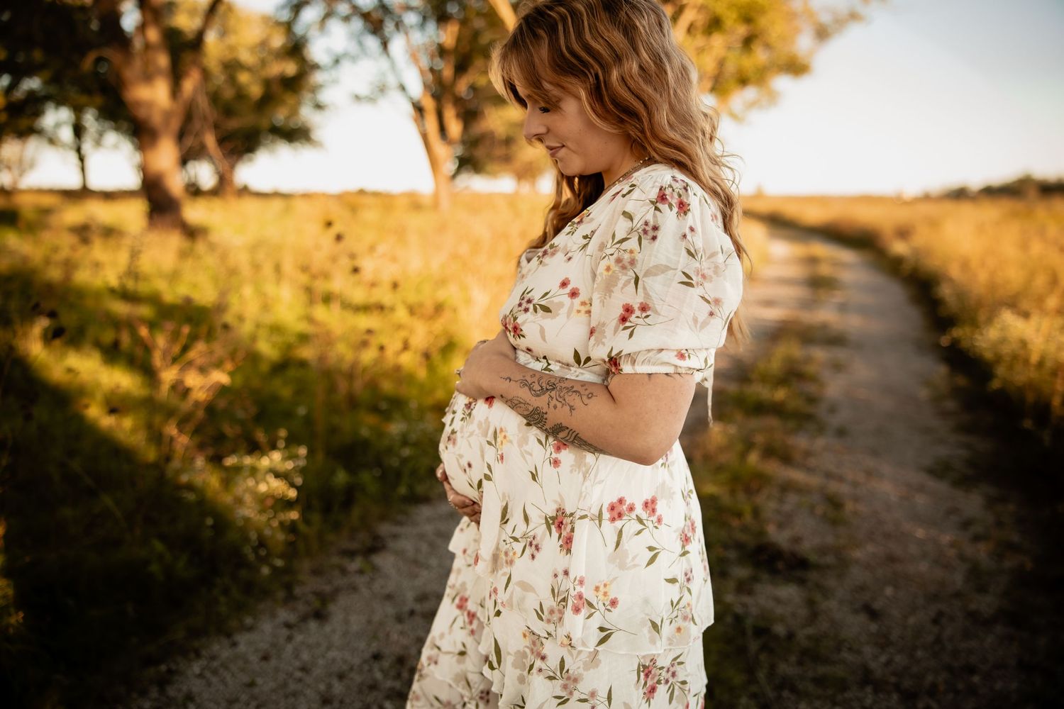 Pregnant woman in floral dress holding baby bump on sunny dirt path in golden hour light.