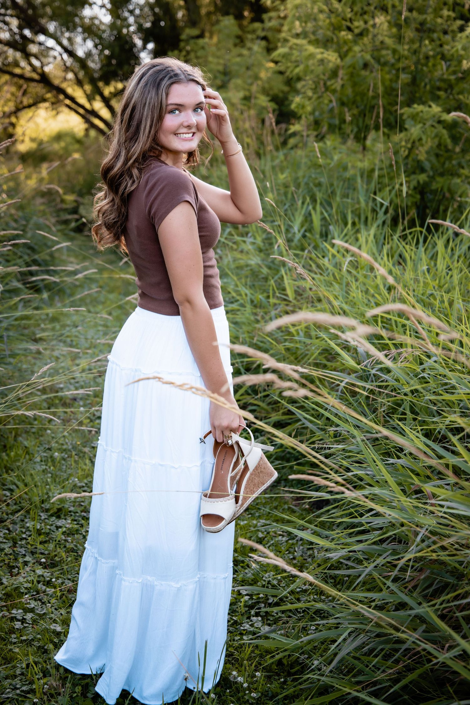 Young woman in white maxi skirt and brown top smiles outdoors, holding sandals among tall green grass.