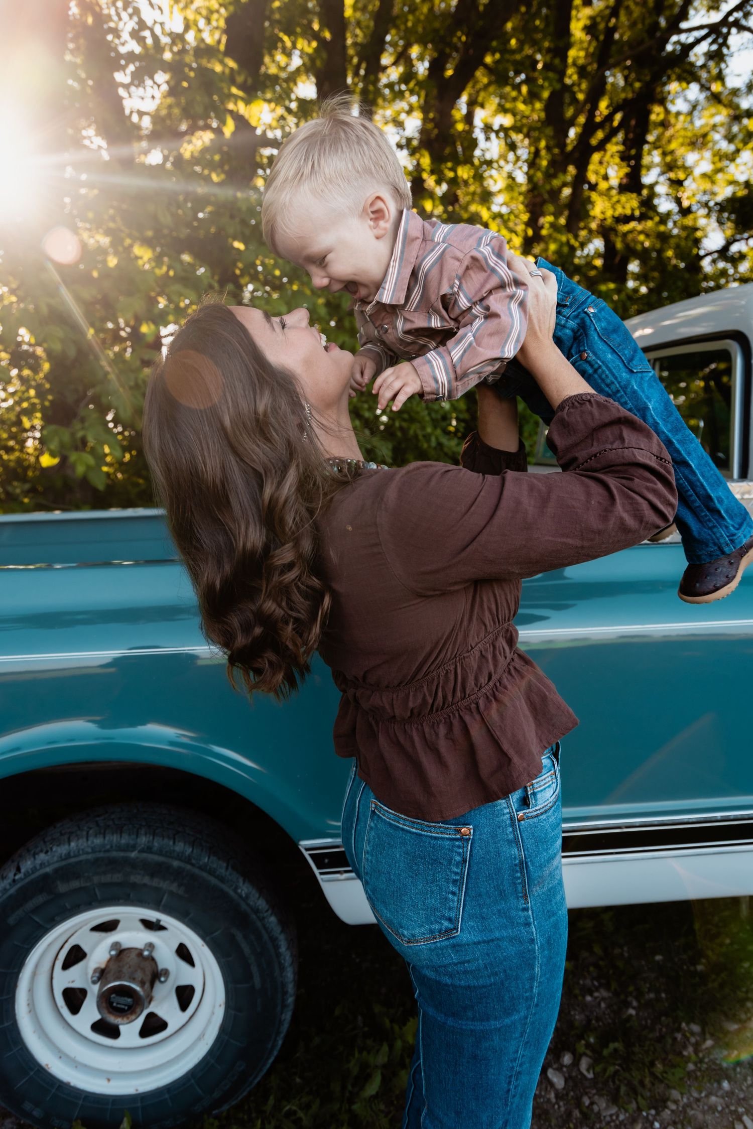 A mother lovingly lifts her toddler son in front of a vintage teal truck in golden sunlight.