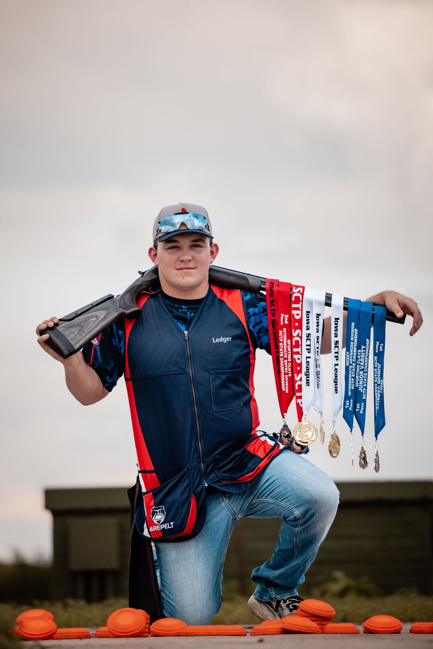 Young male competitive shooter kneeling with shotgun over shoulder displaying multiple SCTP shooting sport medals outdoors.