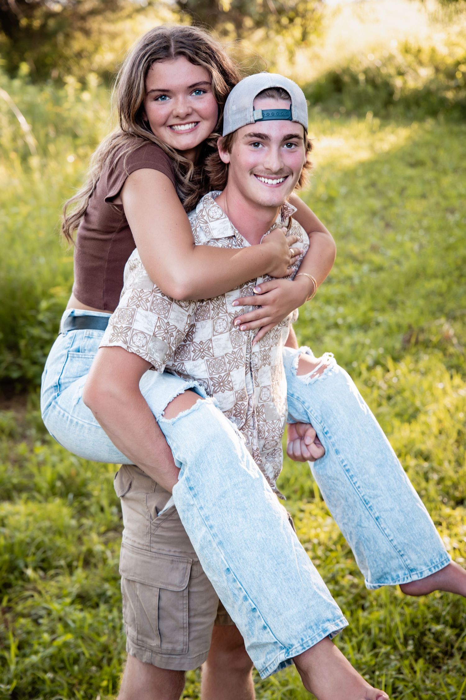 A young couple smiling outdoors, boy giving girl a piggyback ride in a green field.