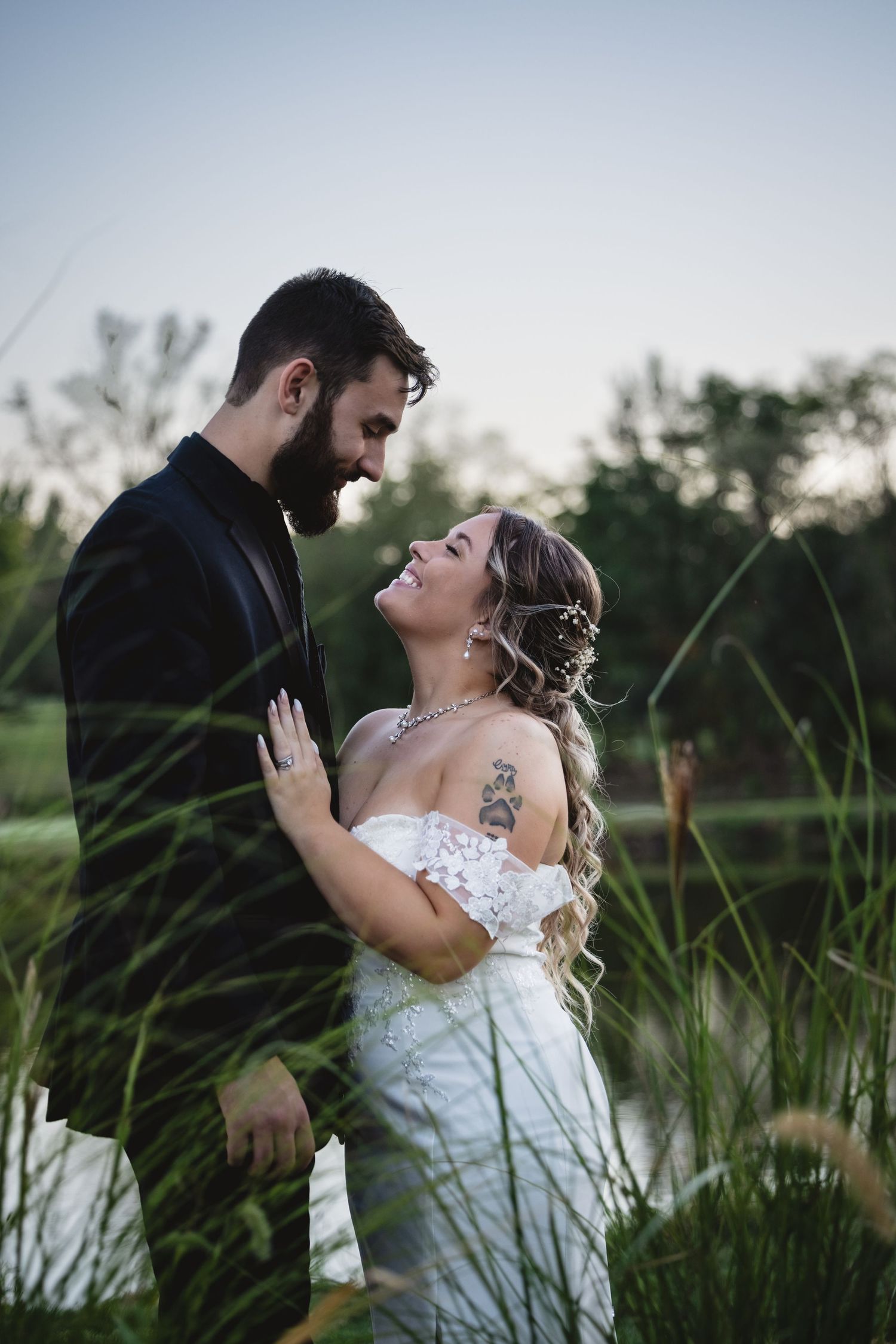 Bride and groom share a joyful moment near tall grasses by a pond at sunset.
