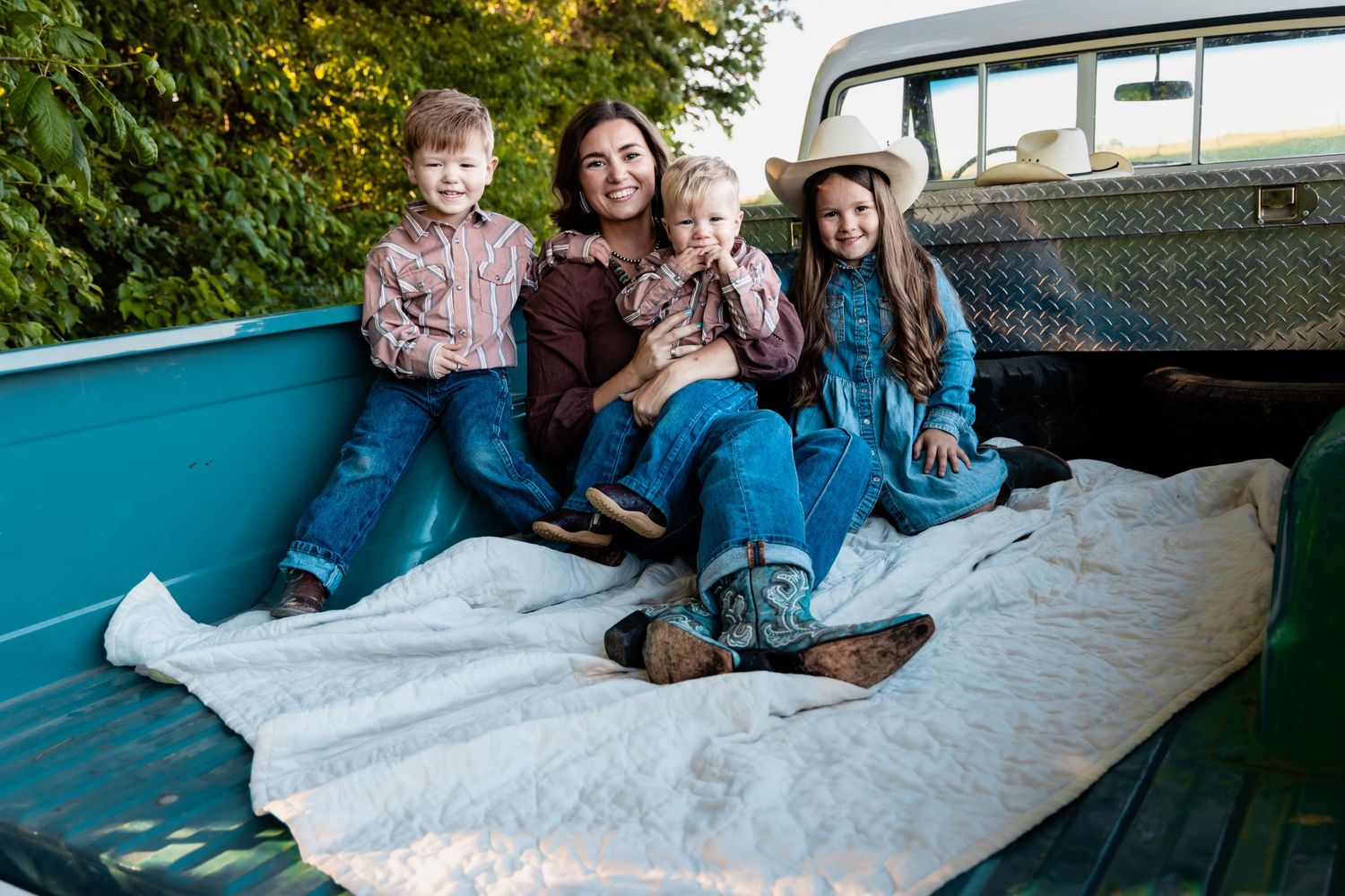 A happy family of four poses together in the bed of a vintage teal pickup truck outdoors.