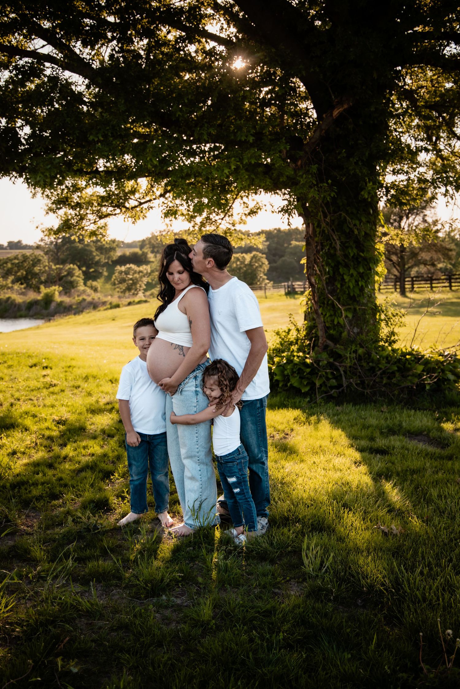 Family maternity photo under large tree at golden hour, parents with two children embracing pregnant mother outdoors.