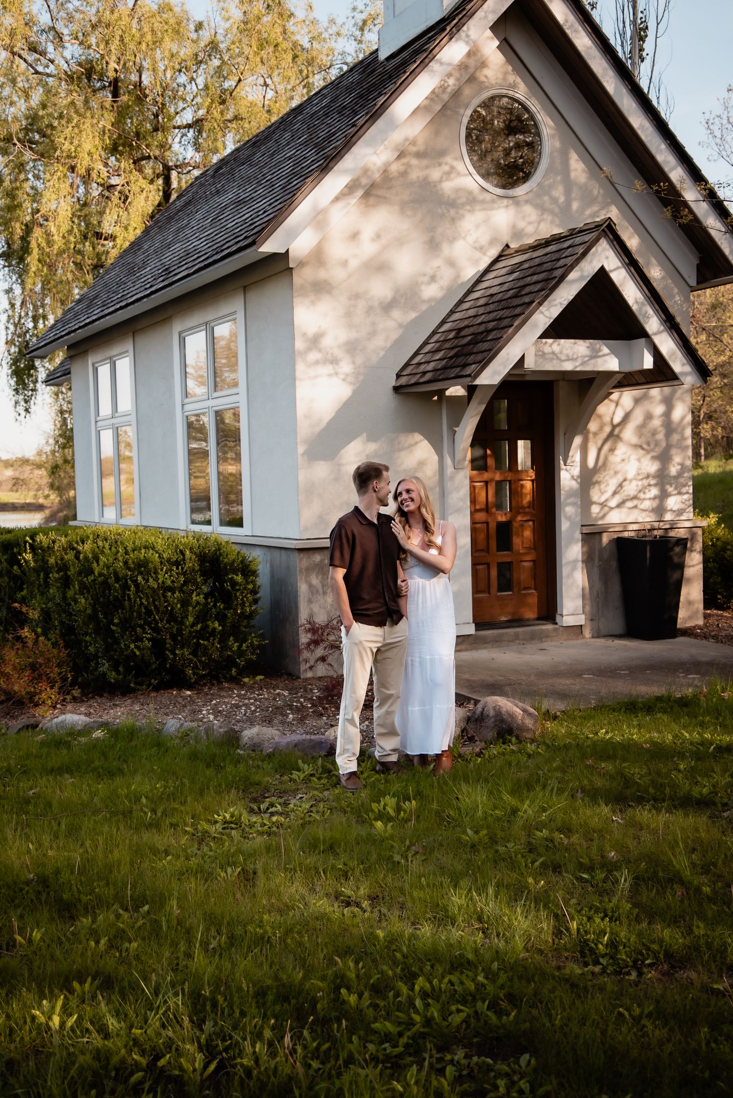 A smiling couple stands together outside a charming small chapel surrounded by lush green landscaping at golden hour.