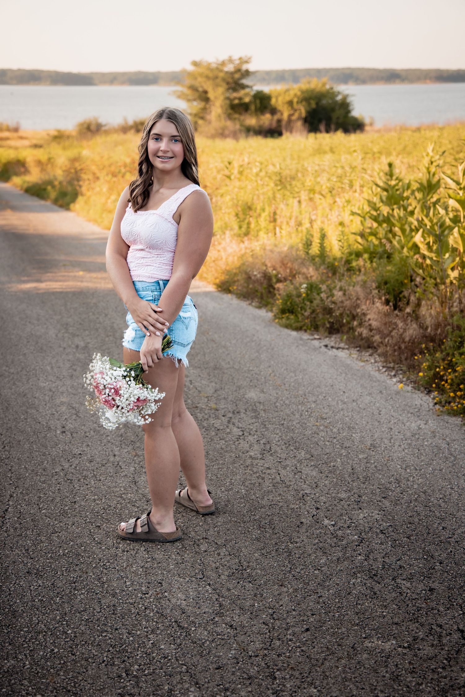 Young woman in denim shorts holding wildflowers on a rural road surrounded by golden fields near a lake.