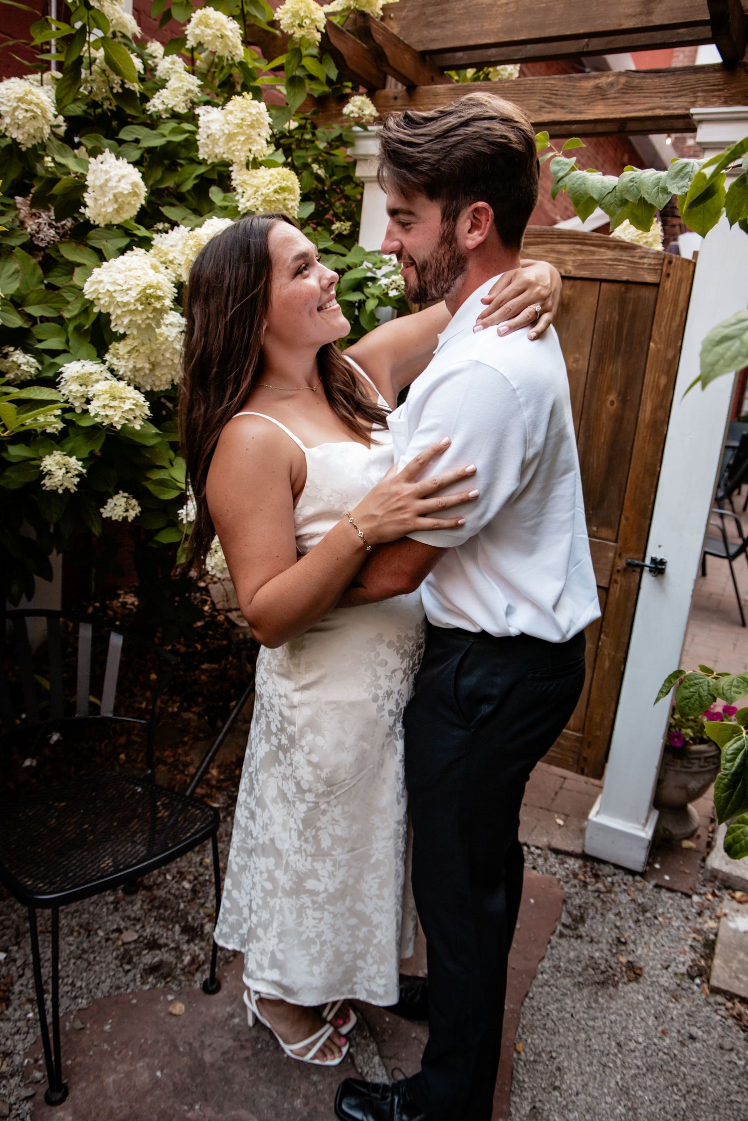 A couple in wedding attire embracing and smiling at each other outdoors near white hydrangeas.