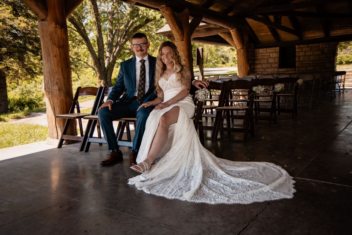Bride in lace gown and groom in blue suit sit together under rustic wooden pavilion at outdoor wedding venue.