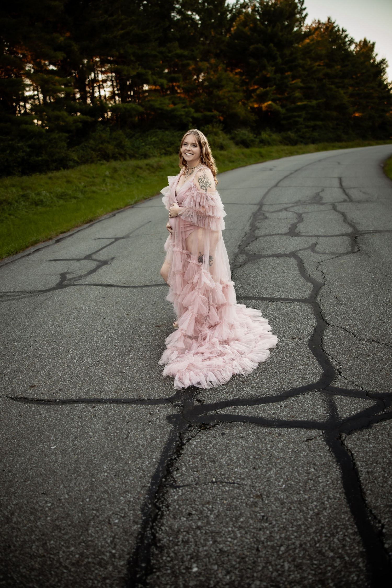 A woman in a flowing pink ruffled gown stands on a cracked road surrounded by lush green trees.