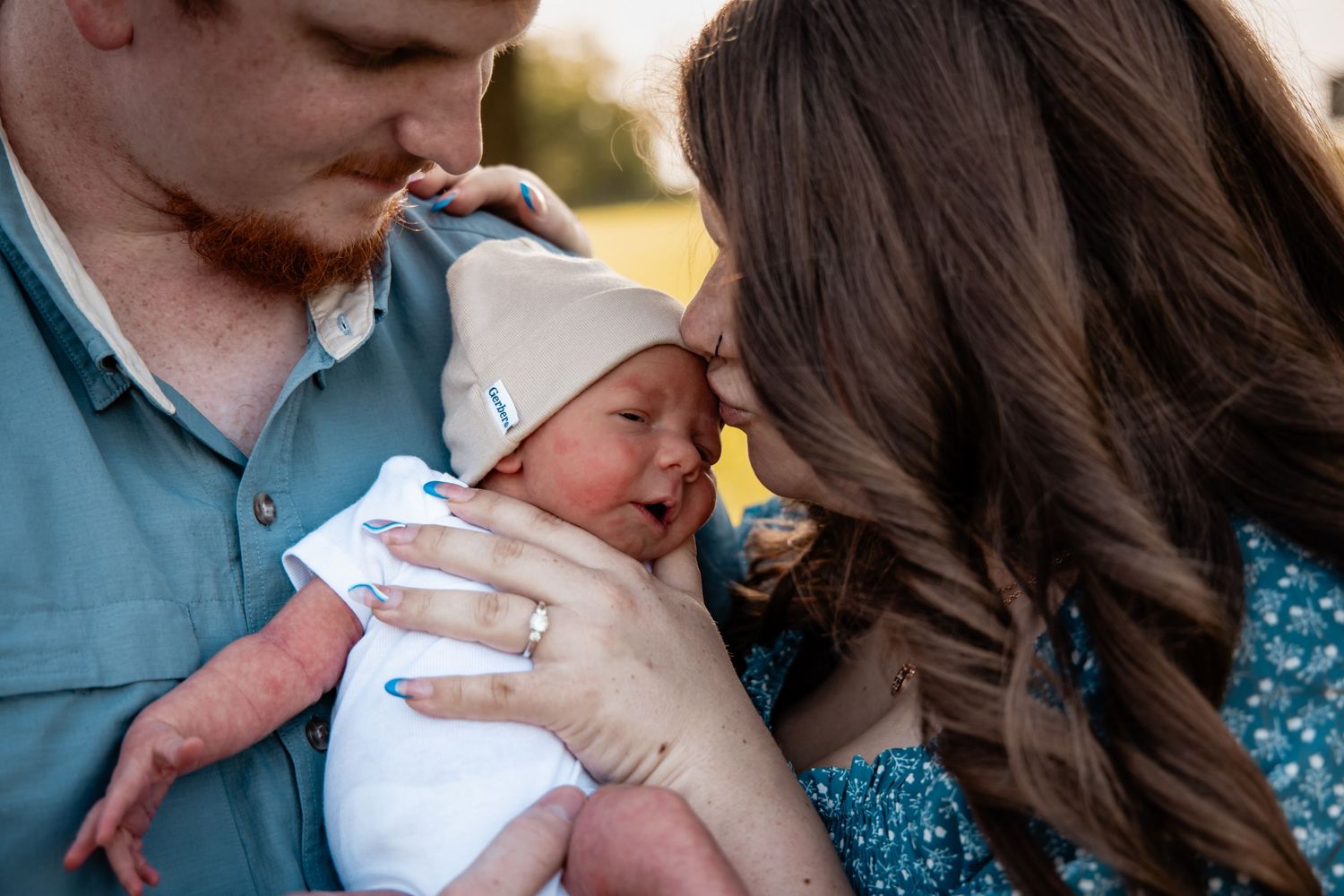 New parents tenderly kissing their swaddled newborn baby outdoors in golden hour light.