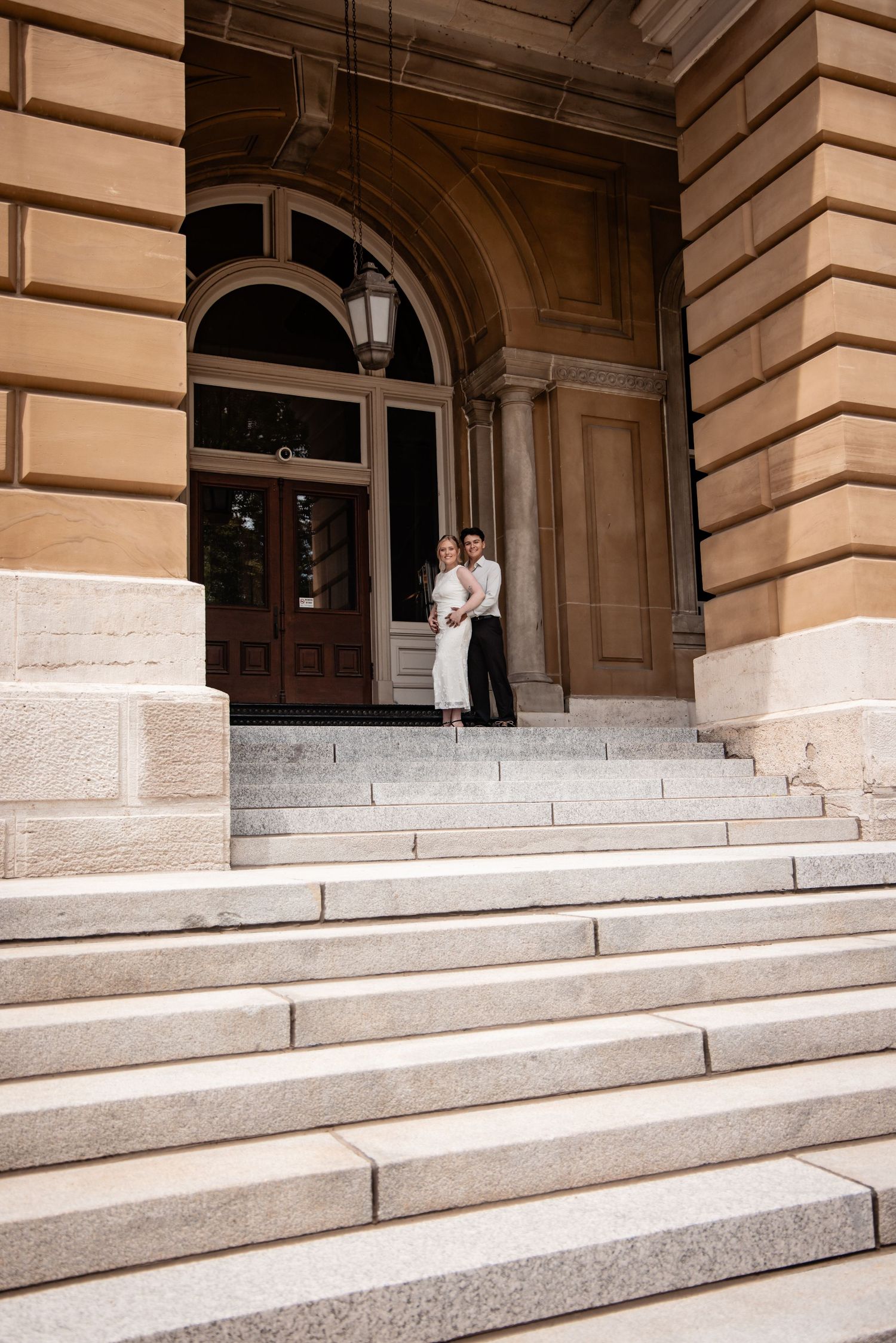 Couple in wedding attire standing on grand stone steps of a classical building with arched entrance.