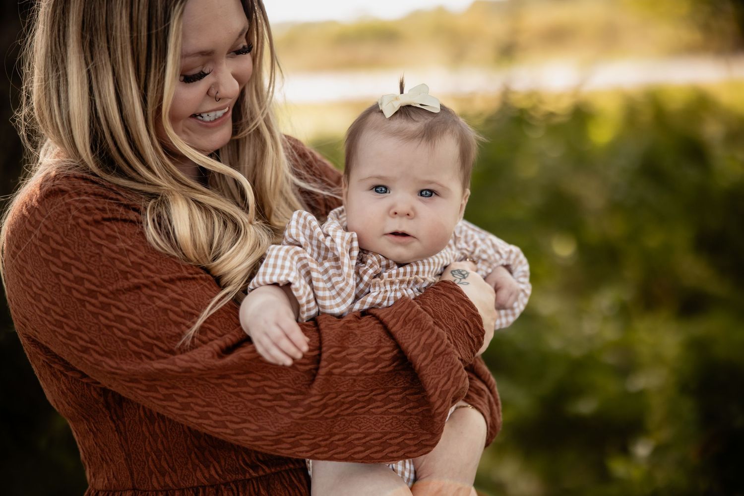 A smiling blonde mother in a rust sweater holds her baby girl wearing a floral outfit and bow outdoors.