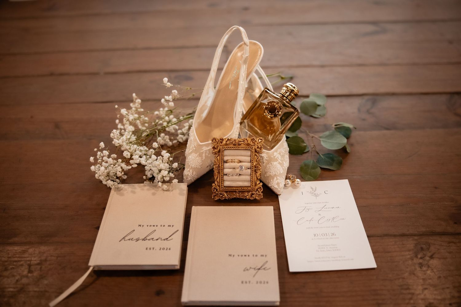 Wedding flat lay with lace bridal shoes, gold perfume bottle, baby's breath flowers, and elegant stationery on wood floor.